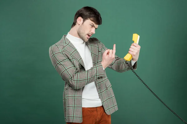 Angry handsome man in vintage clothes showing middle finger in front of retro telephone isolated on green — Stock Photo