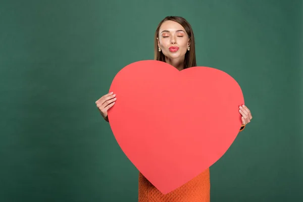 Hermosa mujer haciendo pucheros labios y sosteniendo la tarjeta de papel en forma de corazón con espacio de copia aislado en verde - foto de stock