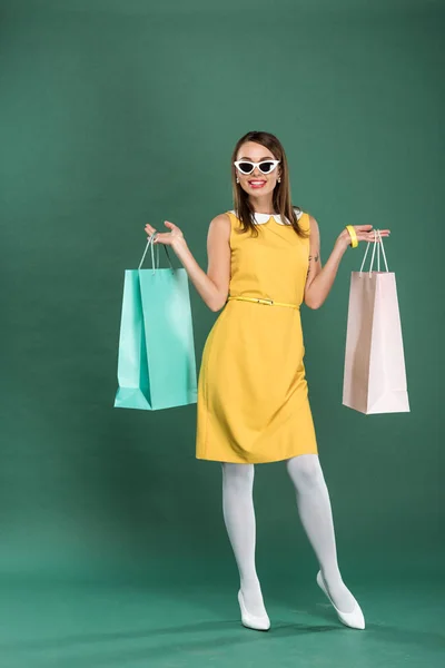 Sorrindo mulher elegante em vestido amarelo e óculos de sol com sacos de compras posando no fundo verde — Fotografia de Stock