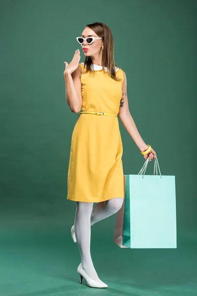 Стильна жінка в жовтій сукні та сонцезахисних окулярах з торговими сумками жестикулює рукою на зеленому фоні — стокове фото