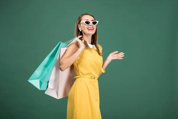 Happy stylish woman in yellow dress and sunglasses with shopping bags isolated on green — Stock Photo