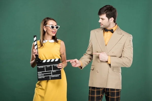 Man in vintage clothes pointing with fingers at woman with clapperboard isolated on green — Stock Photo