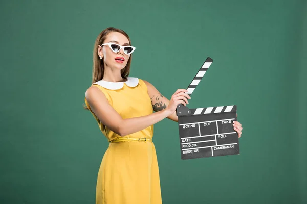 Beautiful stylish woman in yellow dress and sunglasses holding film clapperboard isolated on green — Stock Photo