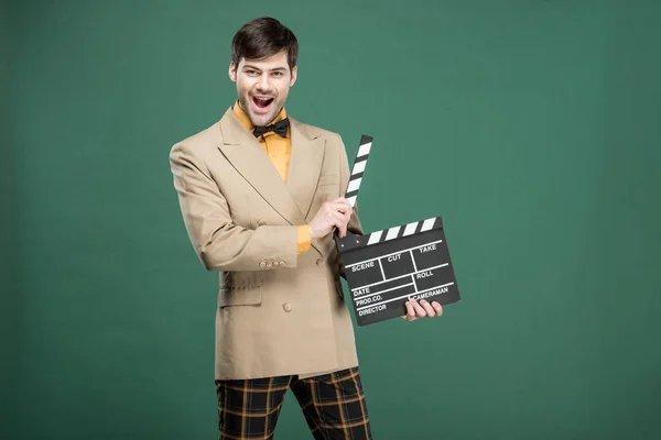 Eccitato bello uomo in abiti vintage guardando la fotocamera e tenendo pellicola clapperboard isolato su verde — Foto stock