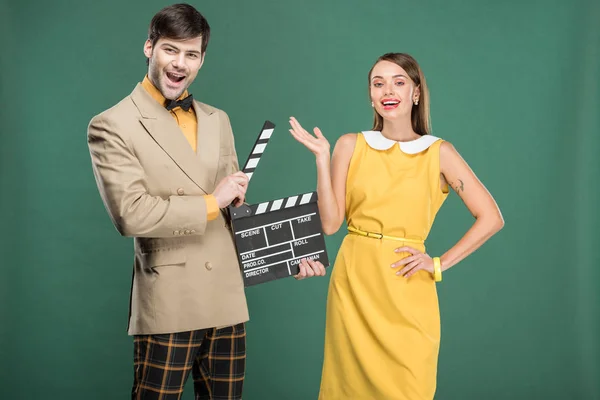 Handsome man in vintage clothes holding film clapperboard while beautiful woman gesturing with hand isolated on green — Stock Photo