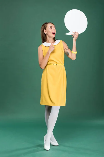 Beautiful stylish woman in yellow dress pointing with finger and holding speech bubble on green background — Stock Photo