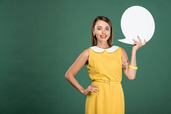 Beautiful smiling woman in yellow dress looking at camera and holding speech bubble isolated on green — Stock Photo