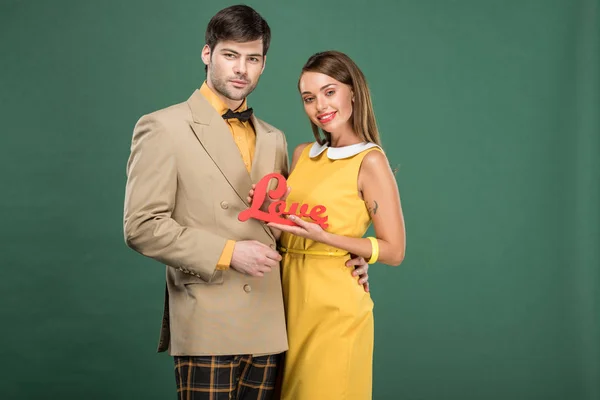 Hermosa pareja en ropa vintage mirando a la cámara y sosteniendo la palabra decorativa 'amor' aislado en verde - foto de stock