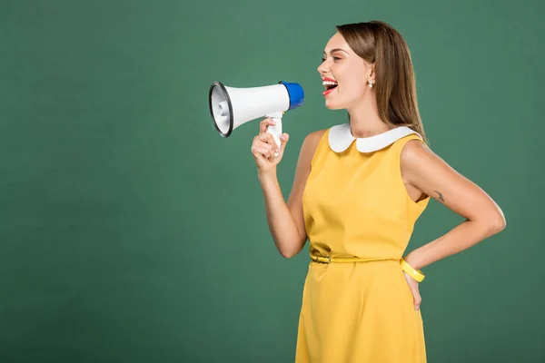 Beautiful stylish woman in yellow dress shouting in loudspeaker isolated on green with copy space — Stock Photo