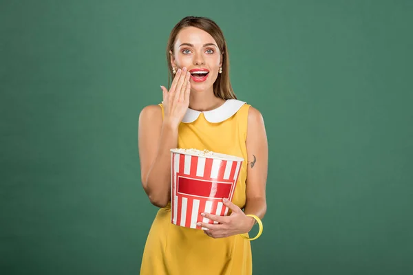Schöne überraschte Frau hält Eimer Popcorn und bedeckt Mund mit Hand isoliert auf grün — Stockfoto
