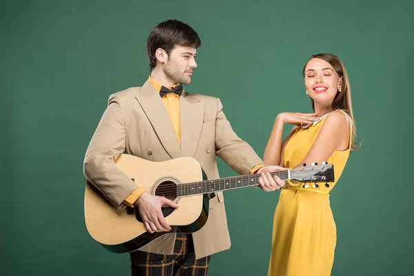 Hombre guapo en ropa vintage tocando la guitarra acústica para hermosa mujer sonriente aislada en verde - foto de stock