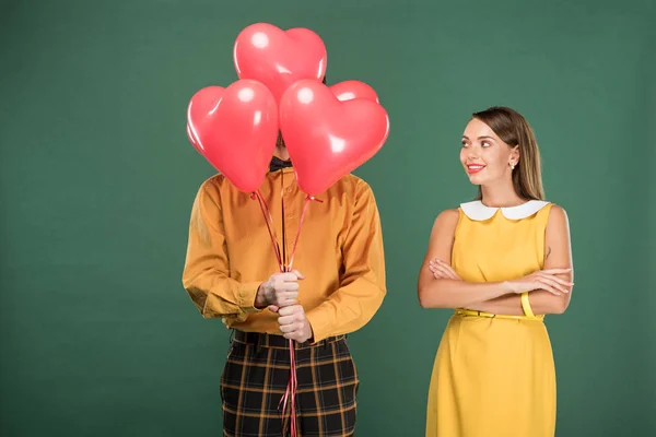 Beautiful woman looking at man covering face with heart shaped balloons isolated on green — Stock Photo