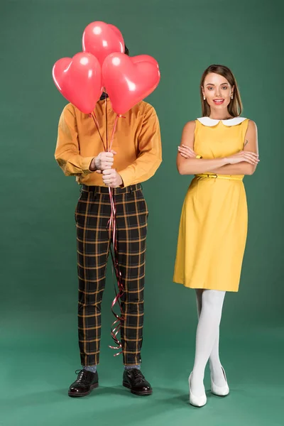 Bela mulher com braços cruzados olhando para a câmera enquanto homem cobrindo rosto com balões em forma de coração no fundo verde — Fotografia de Stock