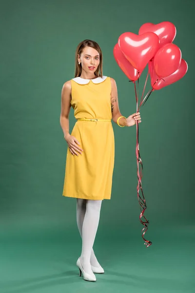 Beautiful woman holding heart shaped balloons and looking at camera on green background — Stock Photo