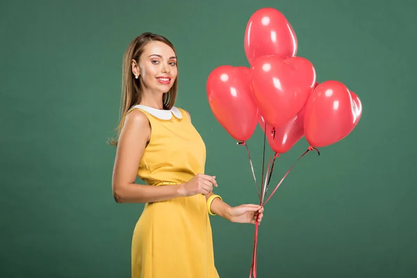 Beautiful woman holding heart shaped balloons and looking at camera isolated on green — Stock Photo