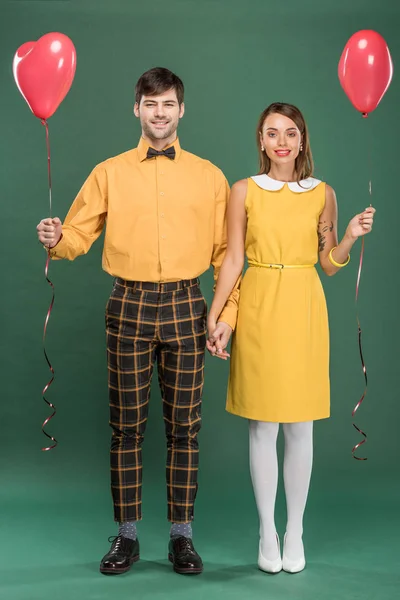 Hermosa pareja sonriente cogida de la mano y globos en forma de corazón mientras mira a la cámara en el fondo verde - foto de stock