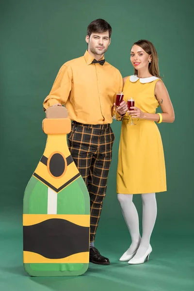 Hermosa pareja en ropa vintage con botella de cartón y copas de vino tinto mirando a la cámara sobre fondo verde - foto de stock