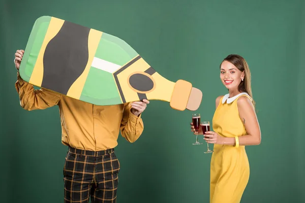 Hombre sosteniendo botella de cartón mientras hermosa mujer en ropa vintage sosteniendo copas de vino tinto aisladas en verde - foto de stock
