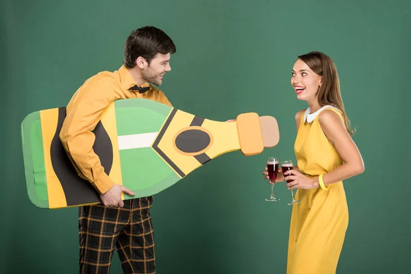 Hermosa pareja en ropa vintage con botella de cartón y copas de vino tinto aisladas en verde - foto de stock
