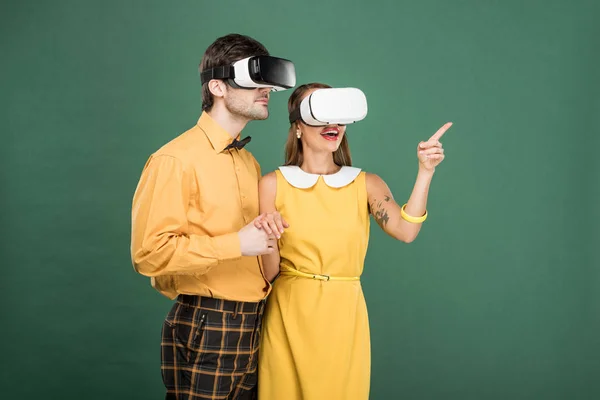 Pareja emocionada en ropa vintage y auriculares de realidad virtual aislados en verde - foto de stock