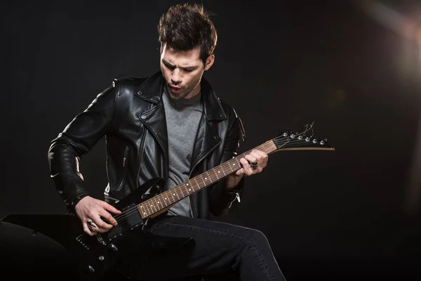Guapo rockero en chaqueta de cuero tocando guitarra eléctrica sobre fondo negro - foto de stock