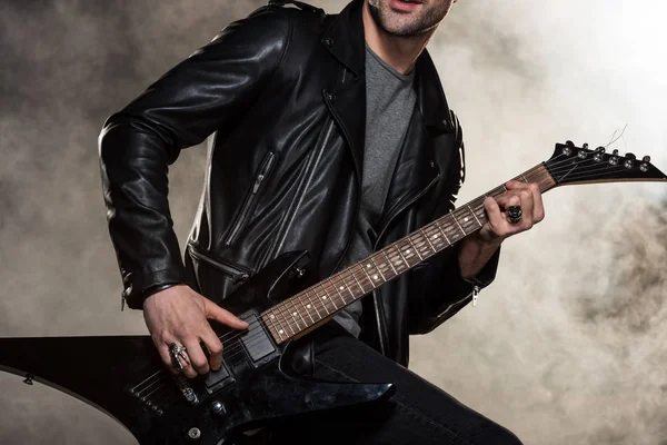 Обрезанный вид рокера в кожаной куртке, играющего на электрогитаре на дымчатом фоне — стоковое фото