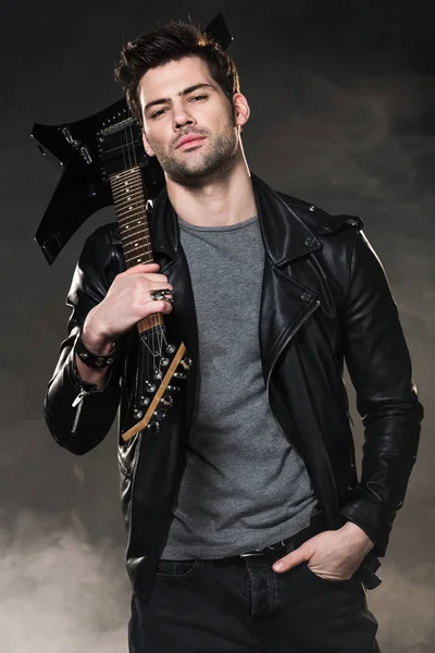 Guapo rockero sosteniendo la guitarra eléctrica y mirando a la cámara sobre fondo oscuro ahumado - foto de stock