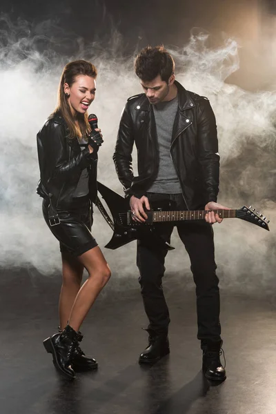 Belle femme en veste en cuir chantant tandis que l'homme jouant de la guitare électrique sur fond fumé — Photo de stock