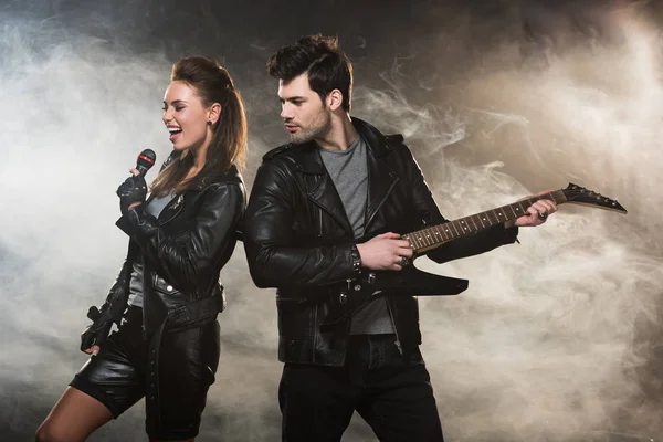 Beautiful woman in leather jacket singing while man playing electric guitar on smoky background — Stock Photo