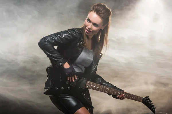 Beautiful woman in leather jacket playing electric guitar on smoky background — Stock Photo