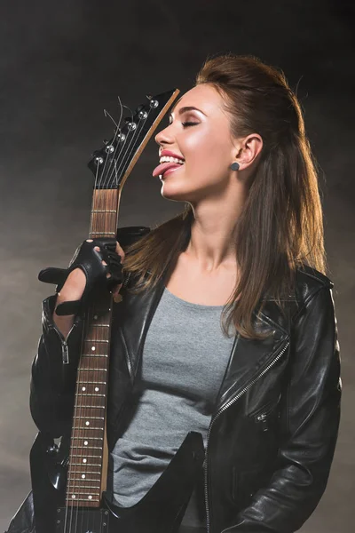 Beautiful woman in leather jacket sticking tongue out and posing with electric guitar on dark background — Stock Photo