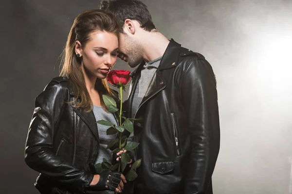 Beau couple en vestes en cuir posant avec rose rouge sur fond fumé — Photo de stock