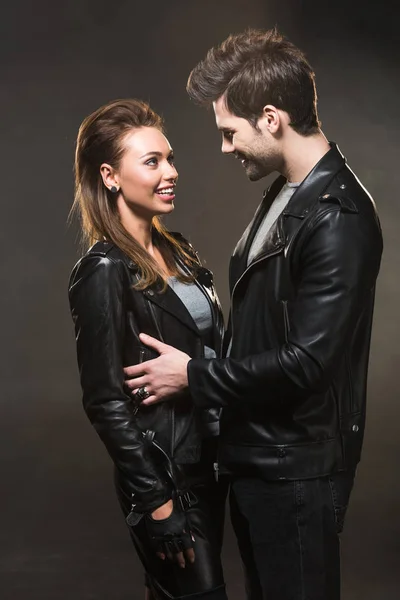 Hermosa pareja sonriente en chaquetas de cuero mirándose el uno al otro y posando sobre fondo oscuro - foto de stock