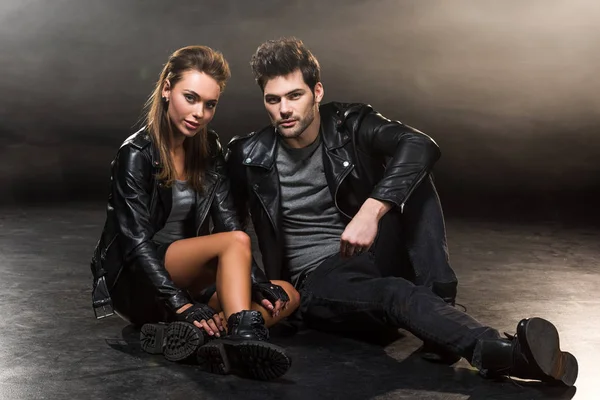 Beautiful fashionable couple in leather jackets sitting and posing on dark background — Stock Photo