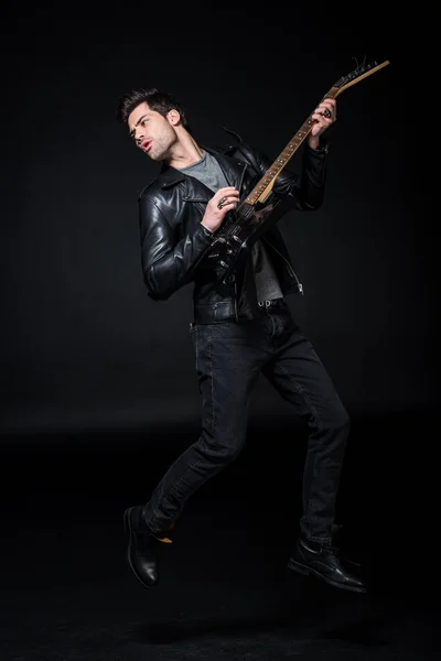 Handsome rocker in leather jacket playing electric guitar isolated on black — Stock Photo