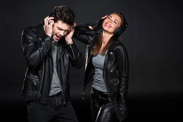 Hermosa pareja en auriculares y chaquetas de cuero escuchando música aislada en negro - foto de stock