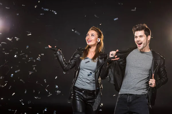 Beautiful happy couple in leather jackets cheering with falling confetti on black background — Stock Photo