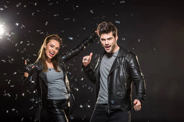 Beau couple en vestes en cuir montrant des signes rock et pouce vers le haut tout en acclamant avec des confettis tombant sur fond noir — Photo de stock