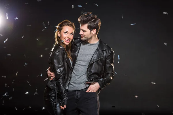 Belo casal em jaquetas de couro abraçando com queda confetti no fundo preto — Fotografia de Stock