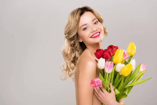Молодая улыбающаяся обнаженная, привлекательная весенняя женщина с букетом разноцветных тюльпанов, изолированных на сером — стоковое фото