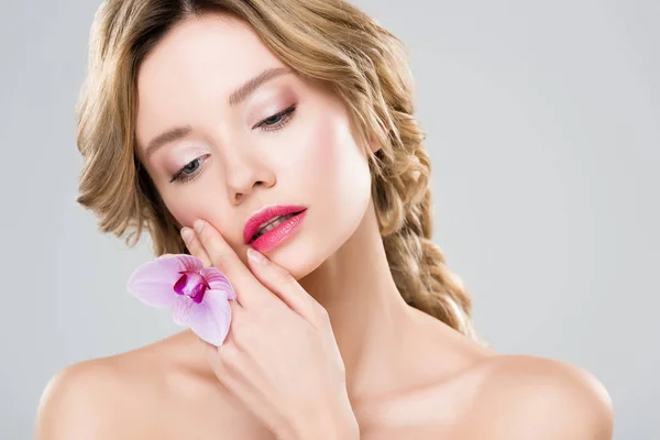 Tierna mujer desnuda tocando la cara con orquídea púrpura en la mano aislado en gris - foto de stock