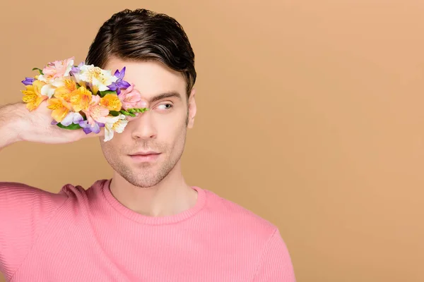 Hombre pensativo que cubre un ojo con flores en la mano aislado en beige - foto de stock