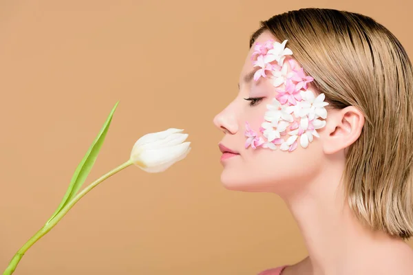 Seitenansicht der eleganten Frau mit Blumen im Gesicht riechende Tulpe isoliert auf beige — Stockfoto