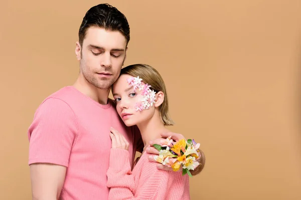 Schöner Mann mit geschlossenen Augen umarmt attraktive Freundin mit Blumen im Gesicht isoliert auf beige — Stockfoto