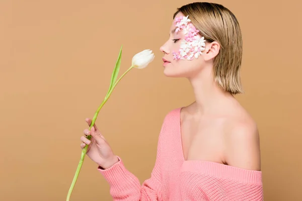 Side view of attractive woman with flowers on face smelling tulip isolated on beige — Stock Photo