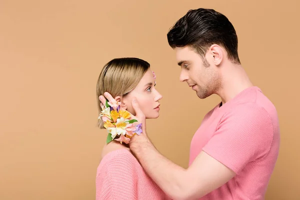 Vista lateral del hombre tierno con flores de alstroemeria en la mano tocando la cara de novia atractiva aislada en beige - foto de stock