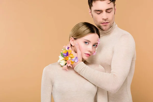Tierno novio con flores de alstroemeria en la mano tocando la cara de hermosa novia aislada en beige - foto de stock
