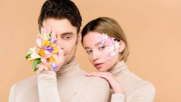Homme avec alstroemeria fleurs à la main couvrant un oeil près de belle petite amie avec des fleurs sur le visage isolé sur beige — Photo de stock