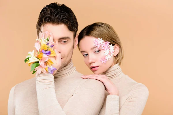 Mann mit Alstroemeria-Blüten an der Hand, die ein Auge in der Nähe einer attraktiven Freundin bedecken, mit Blüten im Gesicht isoliert auf beige — Stockfoto