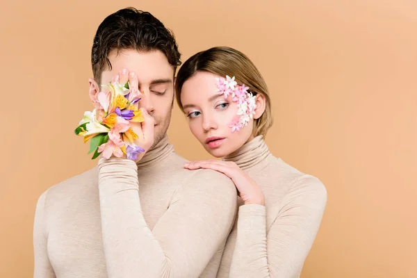 Homme avec des fleurs alstroemeria à la main couvrant un oeil près de belle femme avec des fleurs sur le visage isolé sur beige — Photo de stock